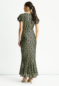 Long, fitted dress in dark green with a metallic pattern, frill cap sleeves, and a flared hemline. Worn with gold heels.
