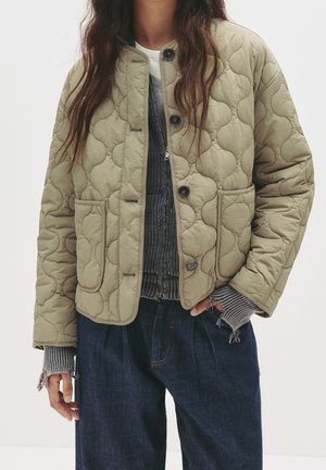 Personne portant une veste matelassée olive clair avec poches avant, un pull gris en maille superposé aux poignets effilochés, et un pantalon en denim bleu foncé.