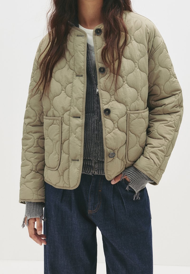 Personne portant une veste matelassée olive clair avec poches avant, un pull gris en maille superposé aux poignets effilochés, et un pantalon en denim bleu foncé.