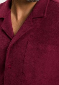 Burgundowa, fakturowana wełniana koszula z dekoltem w kształcie litery V, przednią kieszenią i zapięciem na guziki. Materiał wydaje się miękki i lekko puszysty.