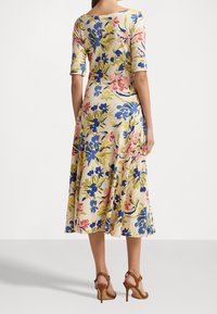 Blommig klänning med trekvartsärm; beige bakgrund med blåa, rosa och gula blommor; utsvängd nederdel; matchad med bruna högklackade skor.