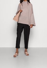 Hellrosa Rollkragenpullover mit geripptem Saum, schwarze, eng zulaufende Hose und rosa Sandalen mit Blockabsatz und Schleifendetail. Beigefarbene Umhängetasche.