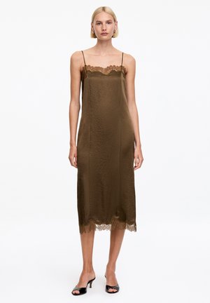 Femme portant une robe nuisette midi vert olive sans manches avec bordure en dentelle et talons ouverts noirs, debout sur un fond blanc.