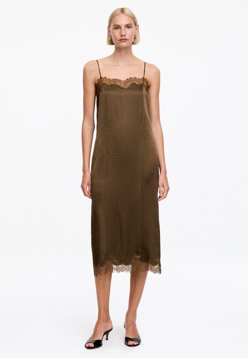 Femme portant une robe nuisette midi vert olive sans manches avec bordure en dentelle et talons ouverts noirs, debout sur un fond blanc.