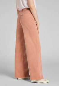 Jeans de pierna ancha, rosa suave, con una cintura alta, que cuentan con bolsillos traseros y detalles de costura sutiles. Combinados con zapatillas blancas.