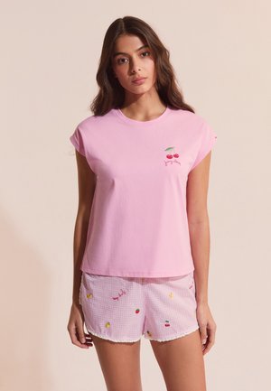 Jeune femme aux longs cheveux bruns portant un haut à manches courtes rose clair avec un logo cerise et un short à carreaux roses avec de petits motifs de fruits.