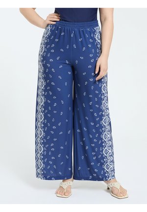 Vrouw draagt blauwe wijde broek met witte paisley- en geometrische randpatronen, gecombineerd met beige sandalen met open tenen, staand met handen in de heupen.