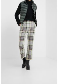 Gepolsterte dunkelgrüne Weste über einem schwarzen Strickpullover, kombiniert mit grau karierter Hose mit rosa und gelben Linien und schwarzen Ankle-Boots.