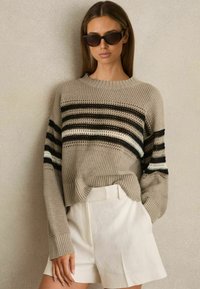 Sweter wełniany w beżowym kolorze z czarno-białymi poziomymi paskami, charakteryzujący się luźnym krojem, ściągaczami przy rękawach i zaokrąglonym dołem. Połączony z białymi szortami.