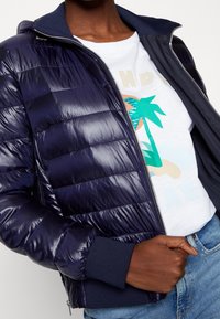 Granatowa kurtka puchowa z połyskującym wykończeniem, poziomym przeszyciem, zapięciem na zamek i ściągaczami na rękawach, noszona na białej koszulce z nadrukiem.
