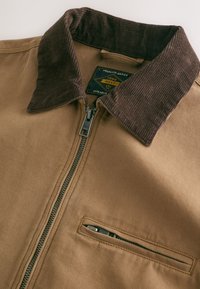 Veste marron clair avec col en velours côtelé marron foncé, fermeture éclair à l'avant et petite poche poitrine zippée, avec une étiquette indiquant "Premium Grade 1982."