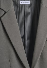 Blazer gris con solapas estructuradas. El interior presenta un forro a rayas en gris oscuro. Etiqueta visible con el nombre de la marca "ANTONY MORATO."