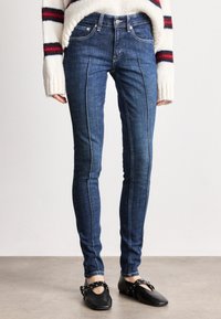Jeans skinny em denim escuro, com costuras em contraste e uma cintura de altura média, combinados com sapatos planos pretos.
