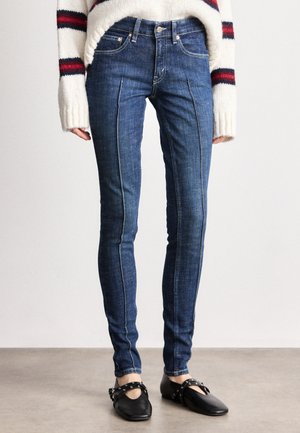 Jean skinny en denim foncé, avec des coutures contrastées et une taille mi-haute, associés à des chaussures noires plates.