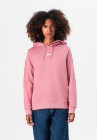 Sweat-shirt rose avec une poche à l'avant, cordon de serrage réglable et détail de logo. Porté avec un jean bleu, il présente une coupe décontractée et des poignets côtelés.