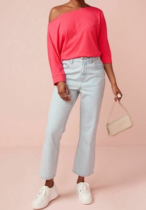 Nainen, jolla on kirkkaan pinkki olkapäät paljastava paita, vaaleansiniset korkeavyötäröiset farkut, valkoiset lenkkarit ja joka pitää pientä beigeä käsilaukkua.