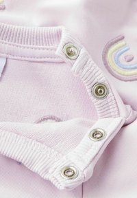 Hele roosa rõivas tekstuurse kangaga, millel on nööpidega kinnitused ja tikandiga vikerkaare detail. Pehme ribane kaelus ja äär.