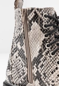 Primer plano de una bota con estampado de piel de serpiente en blanco y negro, que presenta una cremallera beige y cordones negros a través de ojales de metal.