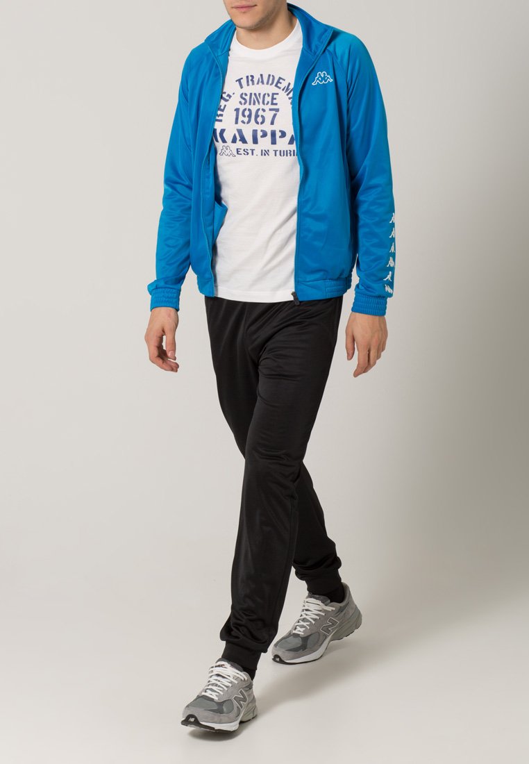 Giacca blu con zip e polsini a costine, t-shirt bianca con grafica e pantaloni sportivi neri. Scarpe da corsa grigie completano il look sportivo.