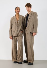 Blazere supradimensionate maro în carouri și pantaloni largi; material texturat cu nasturi întunecați contrasting; asortate cu bluze și pantofi negri.