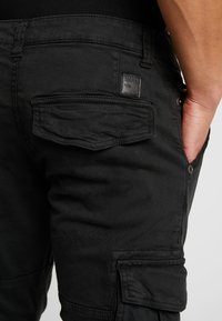 Svarta cargobyxor i bomulldenim, med en bakficka med sömnadsdetalj och en liten märkesetikett. Texturerat tyg med matt yta.