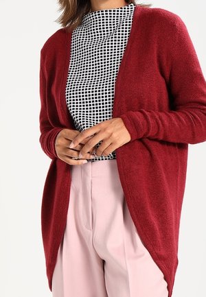 Kobieta ubrana w czerwoną, rozpiętą na przodzie kardigan, czarno-białą bluzkę w kratkę oraz jasnoróżowe, wysokie spodnie, trzymająca ręce splecione przed sobą.