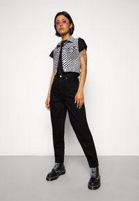 Henkilö, jolla on päällä vaaleanpunaiset aurinkolasit, musta t-paita, mustavalkoinen ruutuvillatakki, mustat korkeavyötäröiset housut ja mustat nauhaliivarikengät, seisoo.