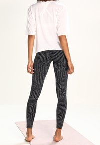 Top blanco de manga corta combinado con leggings negros de cintura alta que presentan un patrón geométrico de cortes con costura blanca sobre un fondo claro.