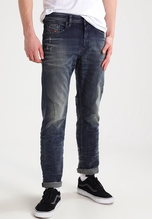 Mężczyzna w wyblakłych, dopasowanych niebieskich jeansach z podwiniętymi nogawkami, białych skarpetkach, czarnych trampkach i białej koszulce na jednolitym tle.