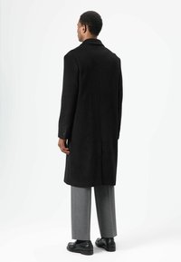 Zwarte wollen overjas met een rechte snit, een getailleerde kraag en met knopen afgewerkte manchetten, gedragen over grijze getailleerde broek en zwarte schoenen.