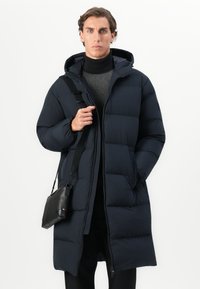 Mand iført en lang, sort hættefrakke over en mønstret sweater, holder en sort crossbody-taskestrop mod en hvid baggrund.