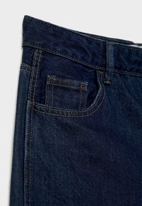 Gros plan d’un jean en denim bleu foncé montrant la poche avant et la petite poche à monnaie avec des surpiqûres jaunes et une passants de ceinture.