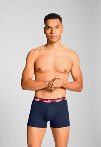 Navy boxer briefs with a red and white Puma logo waistband. The fabric appears smooth, fitted design with a low-rise cut. Model stands with arms folded.