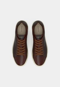 Sneakers din piele maro cu un finisaj neted, vârf rotund, șireturi marron și talpă din cauciuc de culoare crem. Prezintă un branding subtil pe limbă.