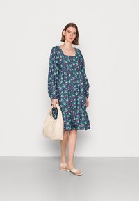 Vestido florido azul-marinho com mangas longas, decote quadrado e saia em camadas; combinado com uma mala bege e sandálias rasas bege claro.