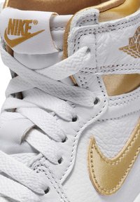 Sneaker alta bianca in pelle con dettagli dorati. Presenta un materiale testurizzato, lacci bianchi piatti e un logo Nike Swoosh dorato.