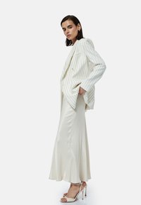 Blazer blanco de rayas, corte relajado, combinado con una elegante falda maxi de satén blanca y sandalias de tacón con tiras, contra un fondo liso.