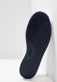 Podešev v černé gumě s vzorovaným gripem, s vyraženým názvem značky. Horní část je bílá s jemnými texturálními detaily.