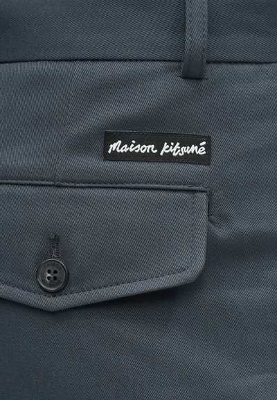 Tmavě šedá látka s hladkým povrchem obsahuje kapsu a černou nášivku s bílým kurzívním textem "maison kitsuné." Viditelné černé knoflíkové detaily.