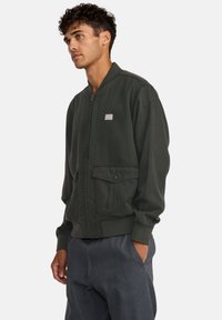 Giubbotto bomber verde scuro con zip, polsini a coste, due tasche frontali e una piccola patch sul petto. Il tessuto appare liscio e resistente.