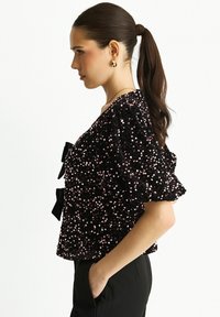 Black sequined top with puffed sleeves and bow accents. The fabric has a textured appearance with pink sequins reflecting light.