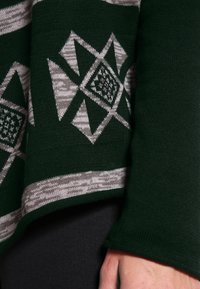 Mörkgrön tröja med ljusgrå geometriska mönster. Har en strukturerad stickning, med en vid, rund halsringning och långa ärmar.