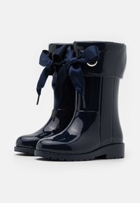Bottes de pluie vernies bleu marine à tige haute, ornées d'un nœud en satin décoratif et d'accents en œillets, sur une semelle noire texturée.