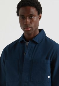 Giovane uomo con capelli ricci corti, che indossa una camicia blu scuro con bottoni e due taschini sul petto, contro uno sfondo bianco.