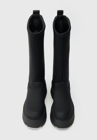 Juodos kelio aukščio batai, pagaminti iš lygios, matinės medžiagos, su masyviu padu, ištraukimo kilputėmis ir minimalistine siuvimo detale palei kraštus.