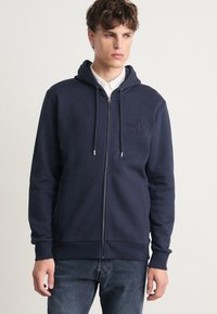 Felpa con zip di colore blu navy realizzata in misto cotone, con cappuccio regolabile tramite cordino e logo in rilievo sul petto. Abbinata a una camicia bianca con colletto sotto.