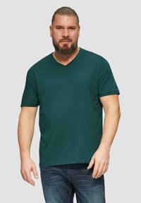 Grünes Baumwoll-V-Ausschnitt-T-Shirt mit kurzen Ärmeln, schlichtem Design und kleinem Logo auf der linken oberen Seite. Getragen mit blauen Jeans.