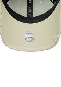 Cappellino da baseball beige con visiera nera. Presenta un patch circolare anteriore che assomiglia a una pallina da baseball. Tessuto texturizzato con bordi sfilacciati.