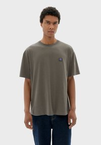 Grijs katoenen T-shirt met korte mouwen, ronde hals en een klein blauw logoplaatje op de linkerborst. Draagt een blauwe spijkerbroek.