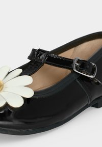 Chaussure Mary Jane en cuir verni noir avec bride à boucle et décoration fleur marguerite blanche sur le devant.